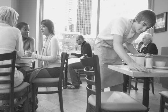 A busser at a coffee shop clears dishes and wipes down a table.