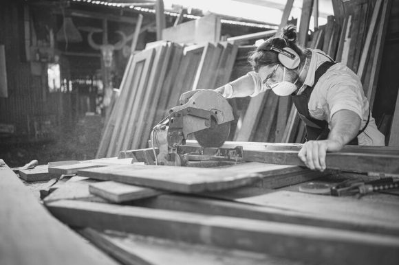 A carpenter, wearing ear and face protection, sawing a board.