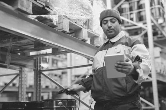 A logistics coordinator stands next to a warehouse shelf, reviewing inventory on a tablet computer.