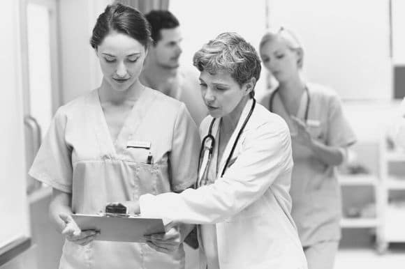 A nurse supervisor, working with her team, points to a clipboard one of her nurses is holding.